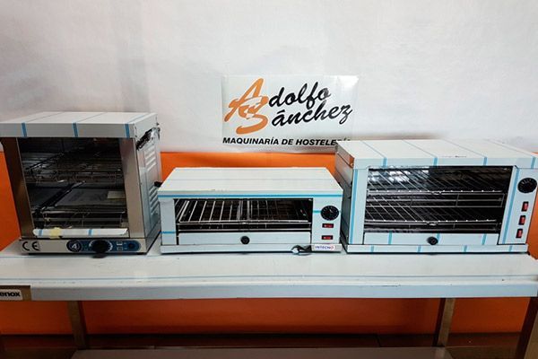 Tres hornos comerciales de acero inoxidable sobre una estantería blanca, con el logo “Adolfo Sánchez” al fondo.