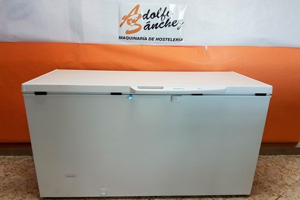 Congelador horizontal de color blanco, con asa, sobre el suelo y con un logotipo al fondo.