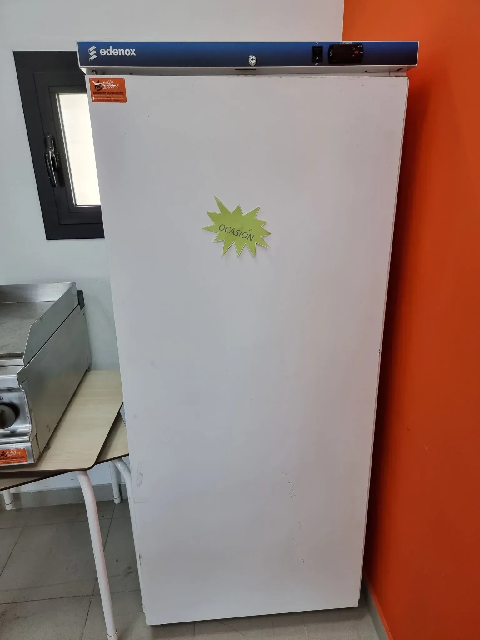 Congelador vertical blanco con tapa azul, contra una pared naranja. Se ven una pequeña ventana y un electrodoméstico de acero inoxidable.