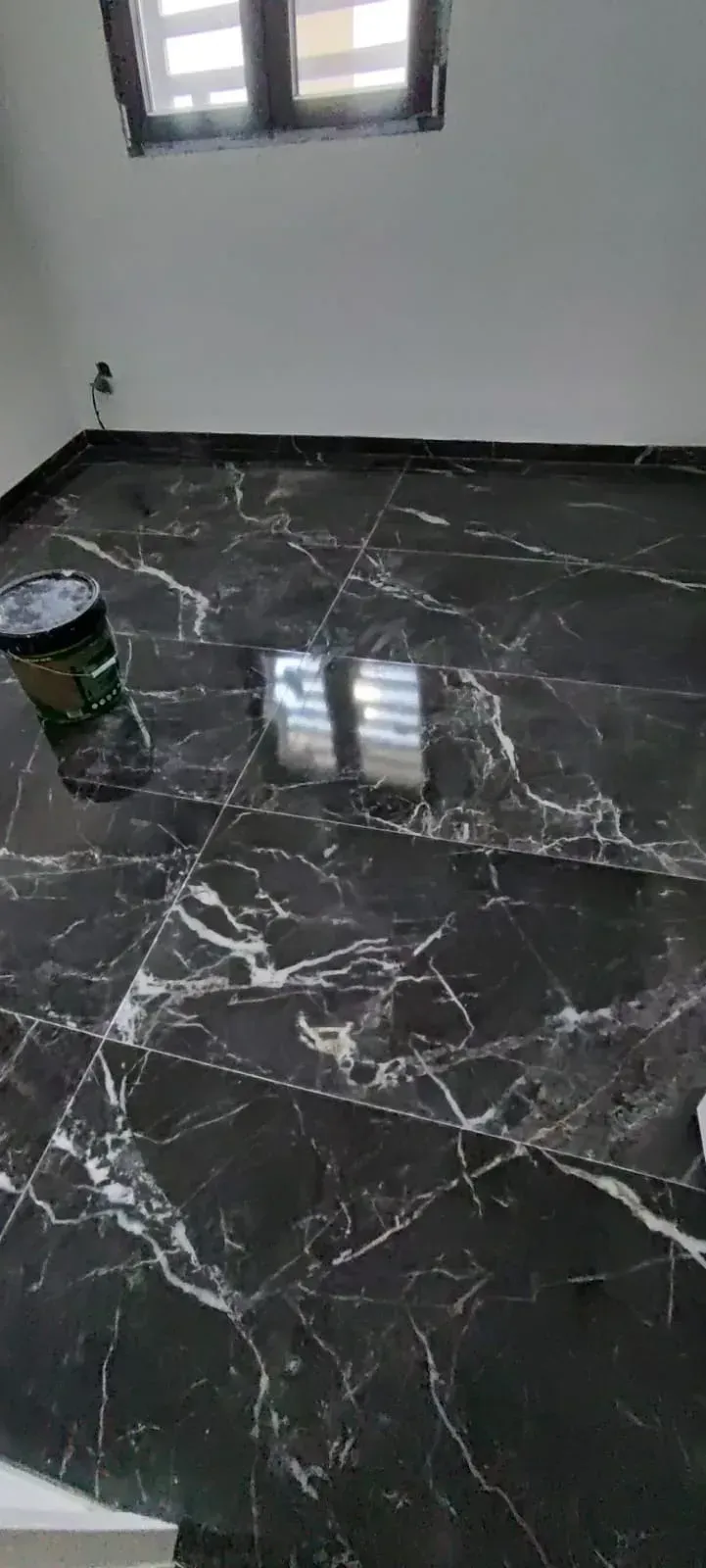 Habitación vacía con suelo de mármol negro brillante, paredes blancas y una pequeña lata de pintura junto a la ventana.