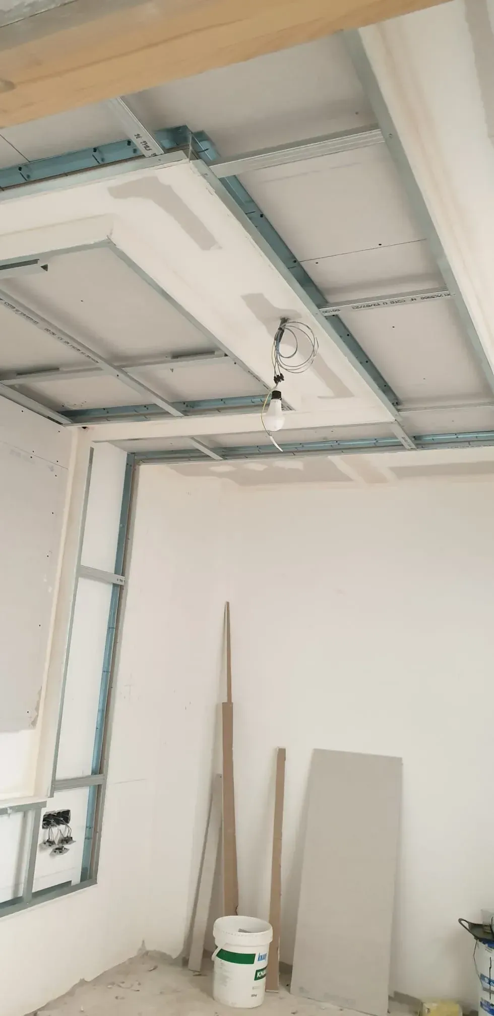 Habitación vacía con paredes blancas, vigas del techo a la vista y materiales de construcción apoyados contra la pared.