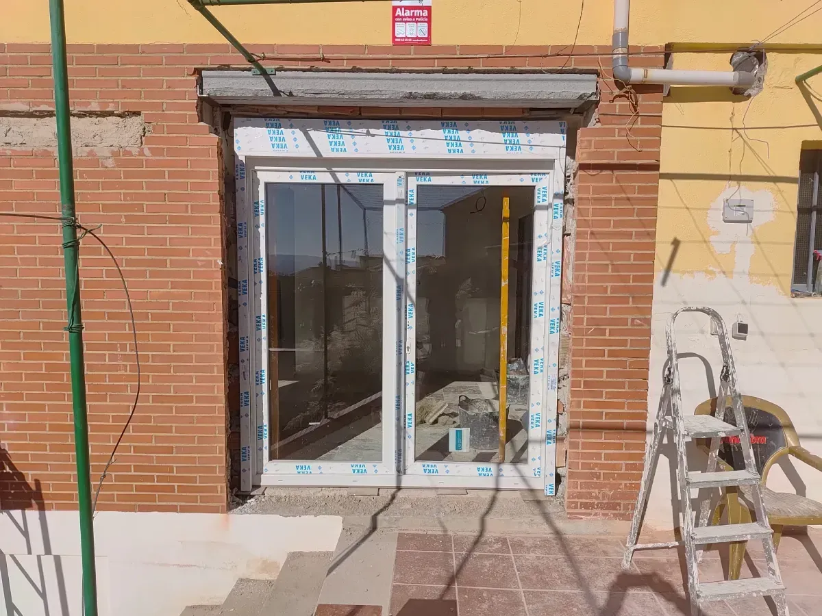 Puerta corredera de cristal en una pared de ladrillo, con una escalera y herramientas de construcción a la derecha.