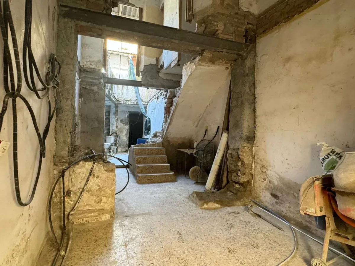 Pasillo interior en ruinas con escaleras, vigas a la vista y herramientas en una habitación polvorienta e inacabada
