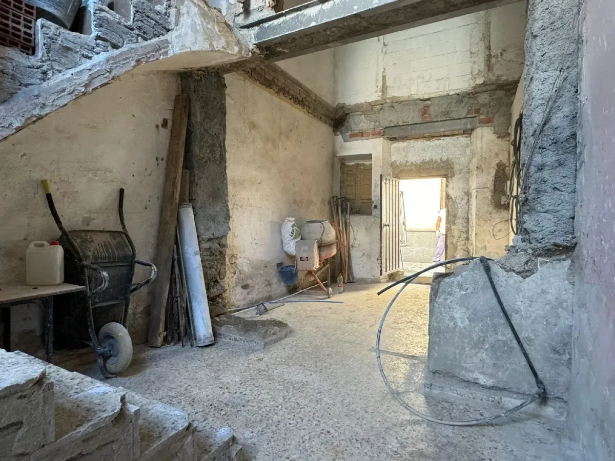 Interior abandonado con paredes de piedra sin revestir, herramientas esparcidas y una puerta luminosa al fondo