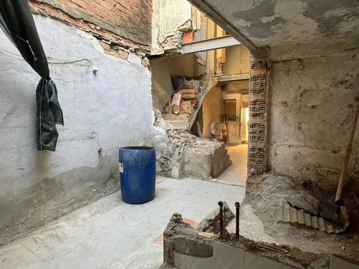 Interior de un sótano en mal estado, con paredes desmoronadas, escombros y un barril azul junto a una abertura