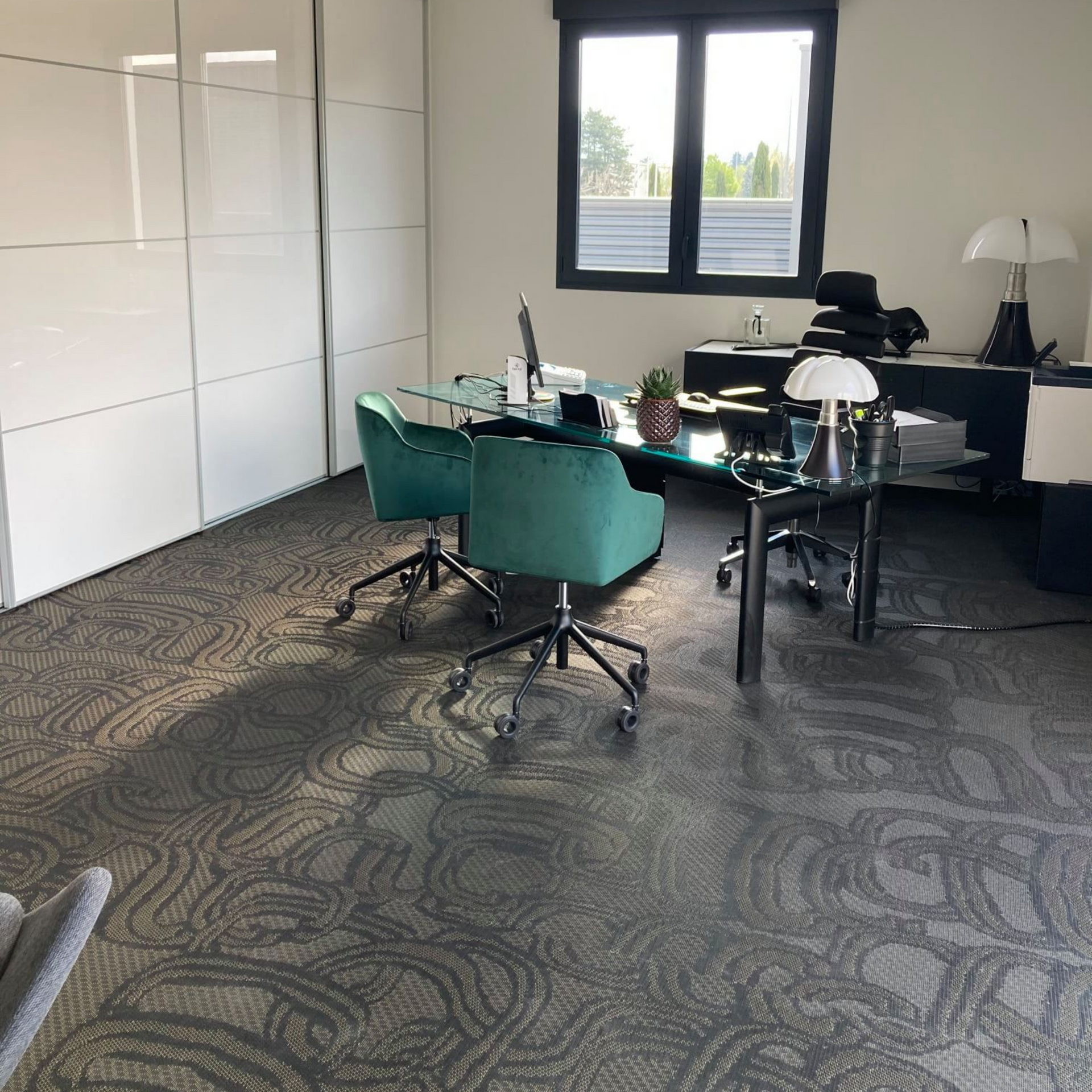 Espace de bureau avec moquette à motifs, chaises turquoise, bureau en verre, armoires blanches et fenêtre.