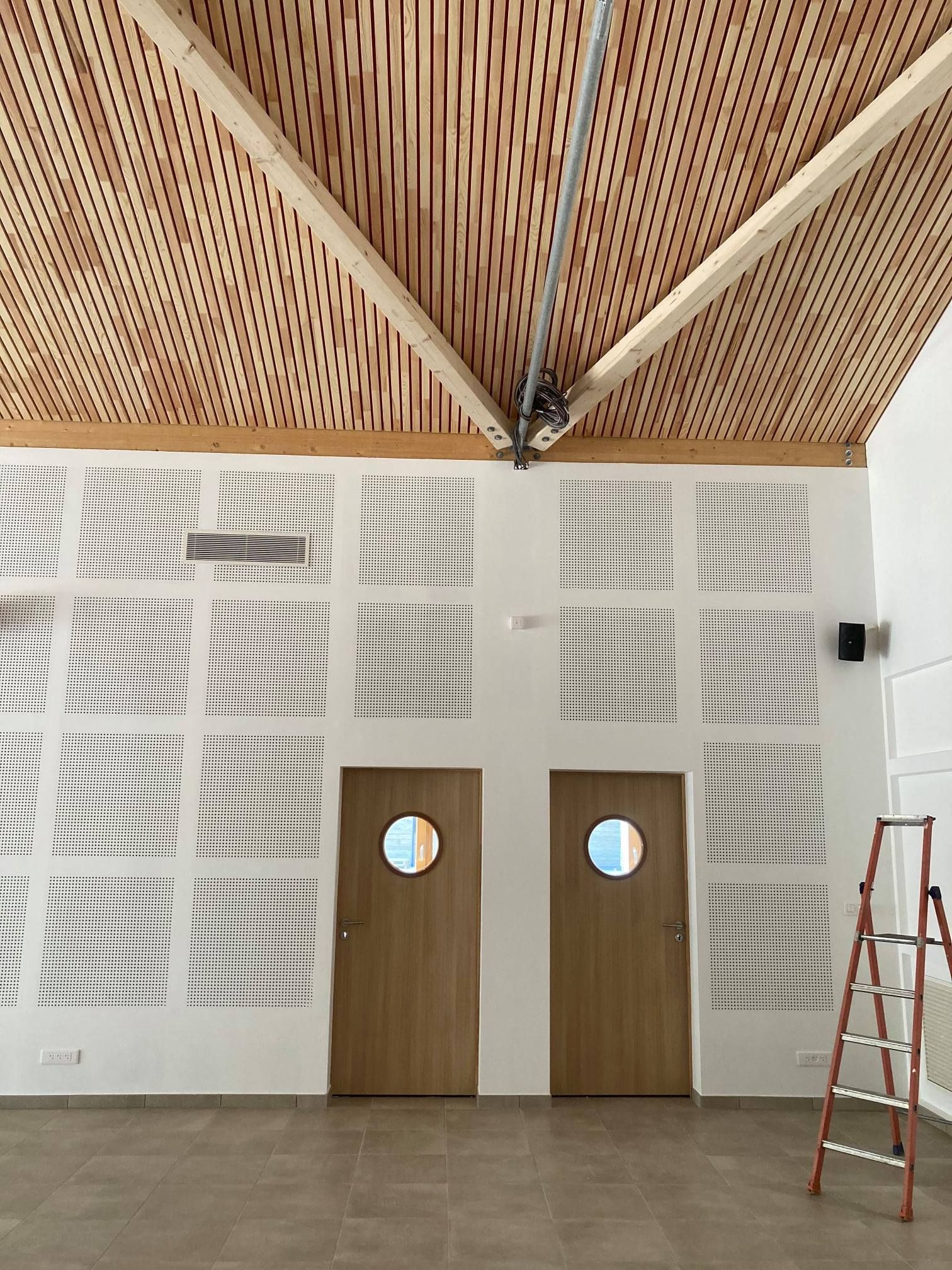 Intérieur d'une pièce avec un plafond à lattes de bois, deux portes et des murs blancs perforés. Une échelle se trouve à droite.