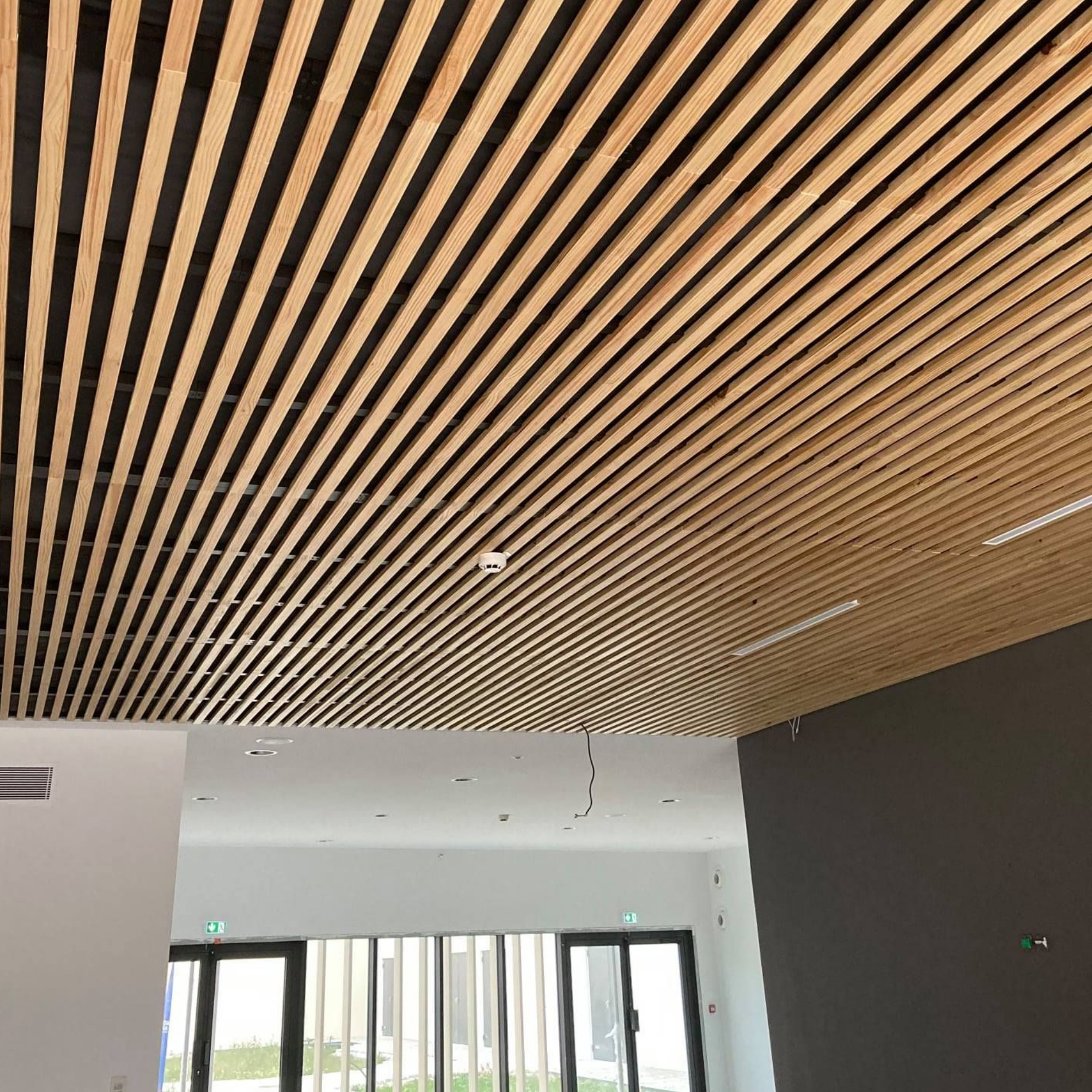 Plafond à lattes de bois avec éclairage encastré au-dessus d'une pièce avec fenêtres et mur gris.