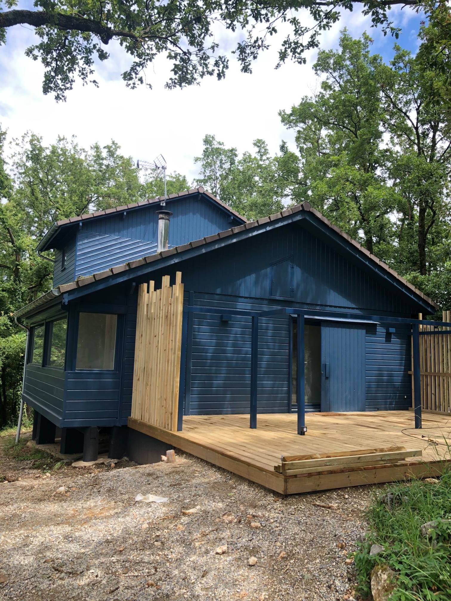Cabane bleue avec terrasse et porche en bois, entourée d'arbres.