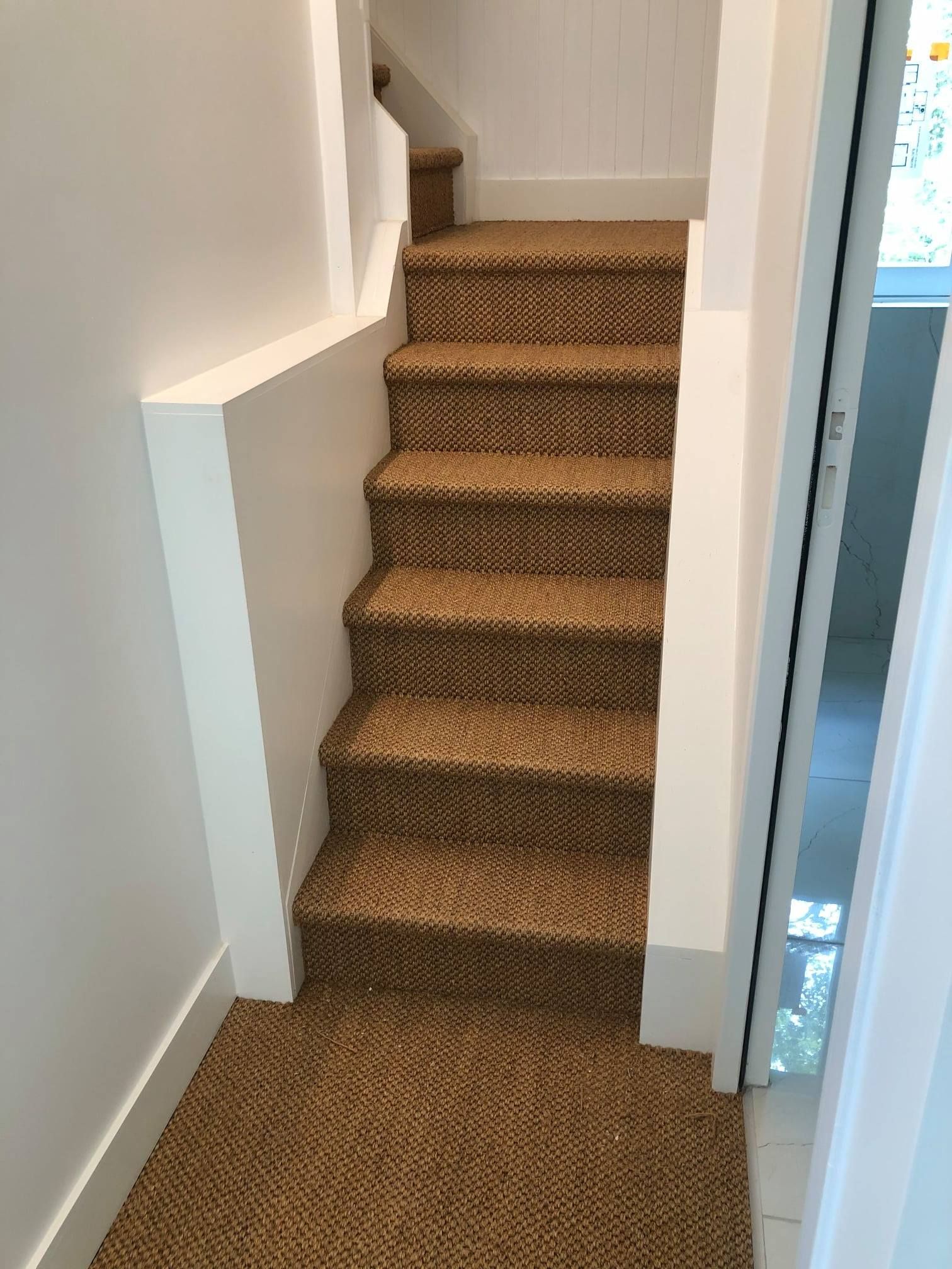 Escalier avec moquette marron et murs blancs. Un cadre de porte se trouve à droite.