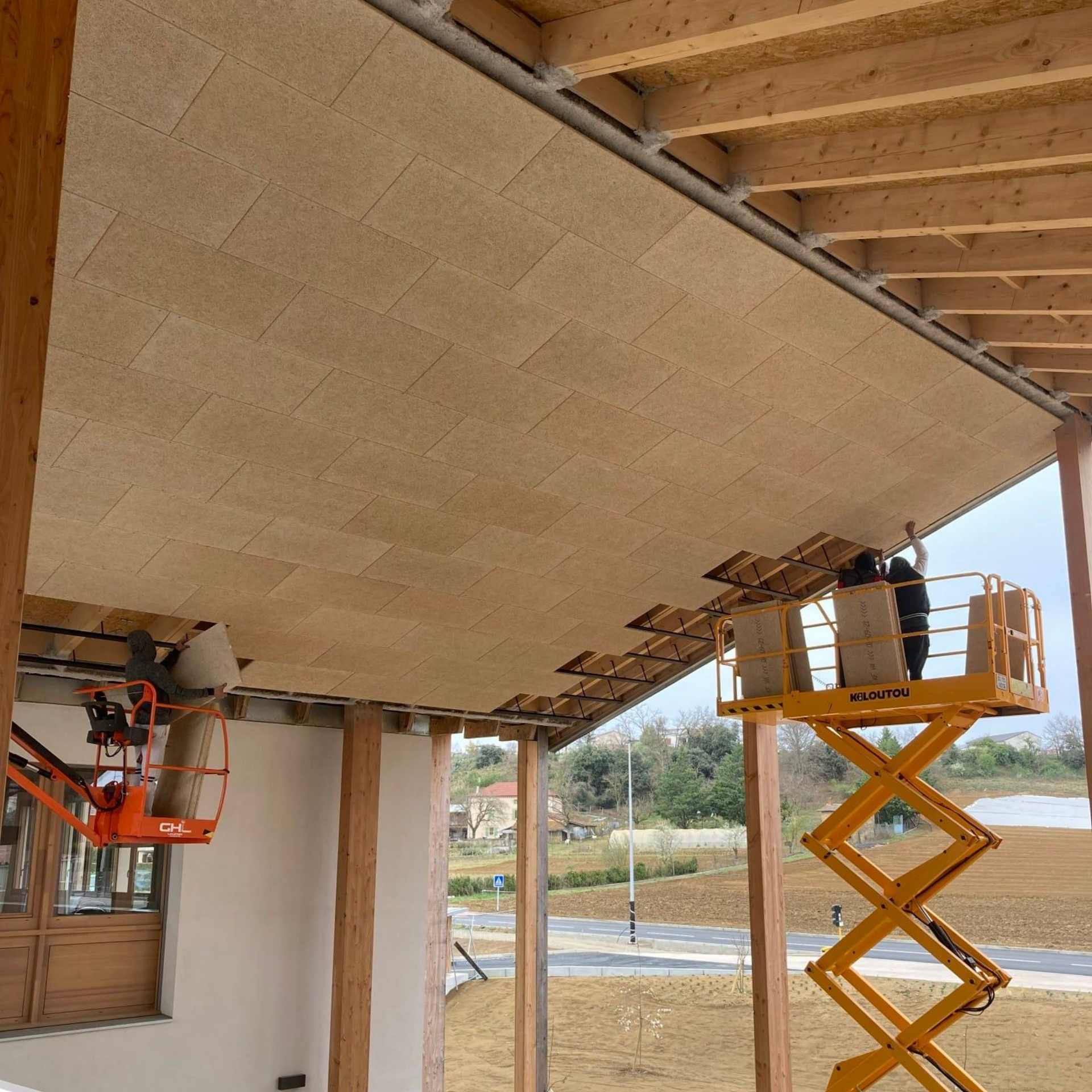 Des ouvriers installent des panneaux sur le plafond d'un bâtiment à l'aide d'une nacelle. Panneaux beiges, poutres en bois et ambiance ensoleillée en extérieur.