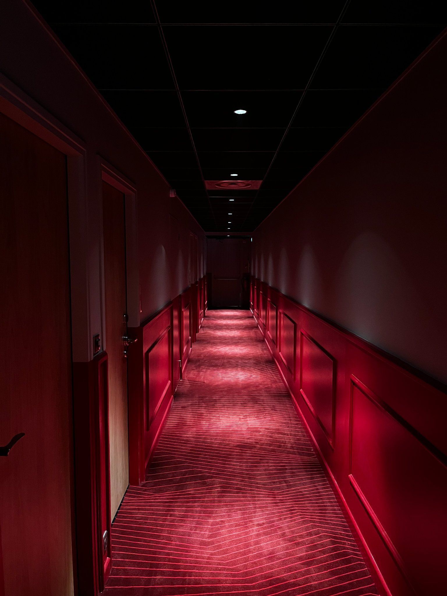 Couloir d'hôtel éclairé en rouge, avec des portes fermées de chaque côté, menant à une porte au loin.
