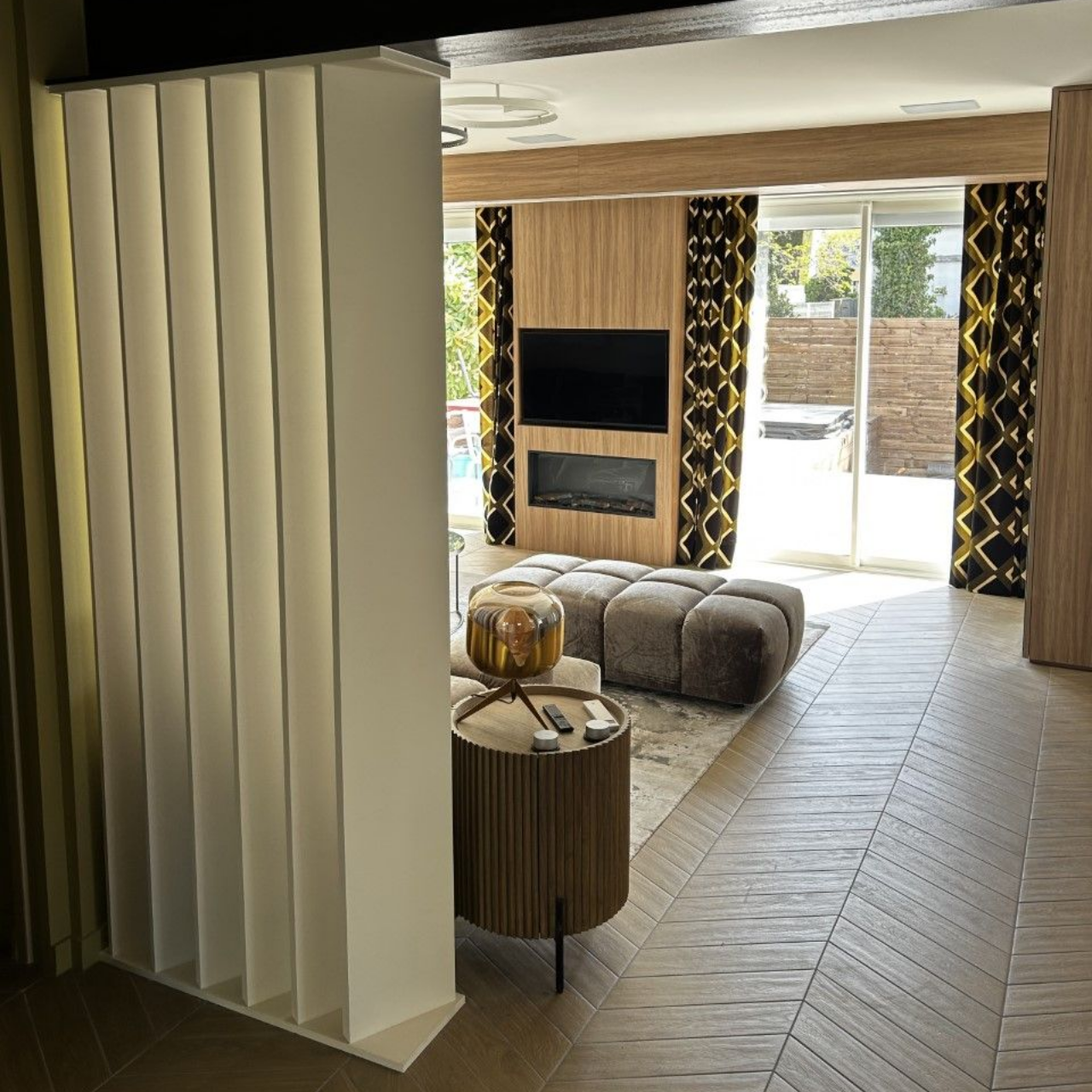 Vue intérieure d'un salon moderne avec une cloison, une télévision et de grandes fenêtres habillées de rideaux noirs et dorés.