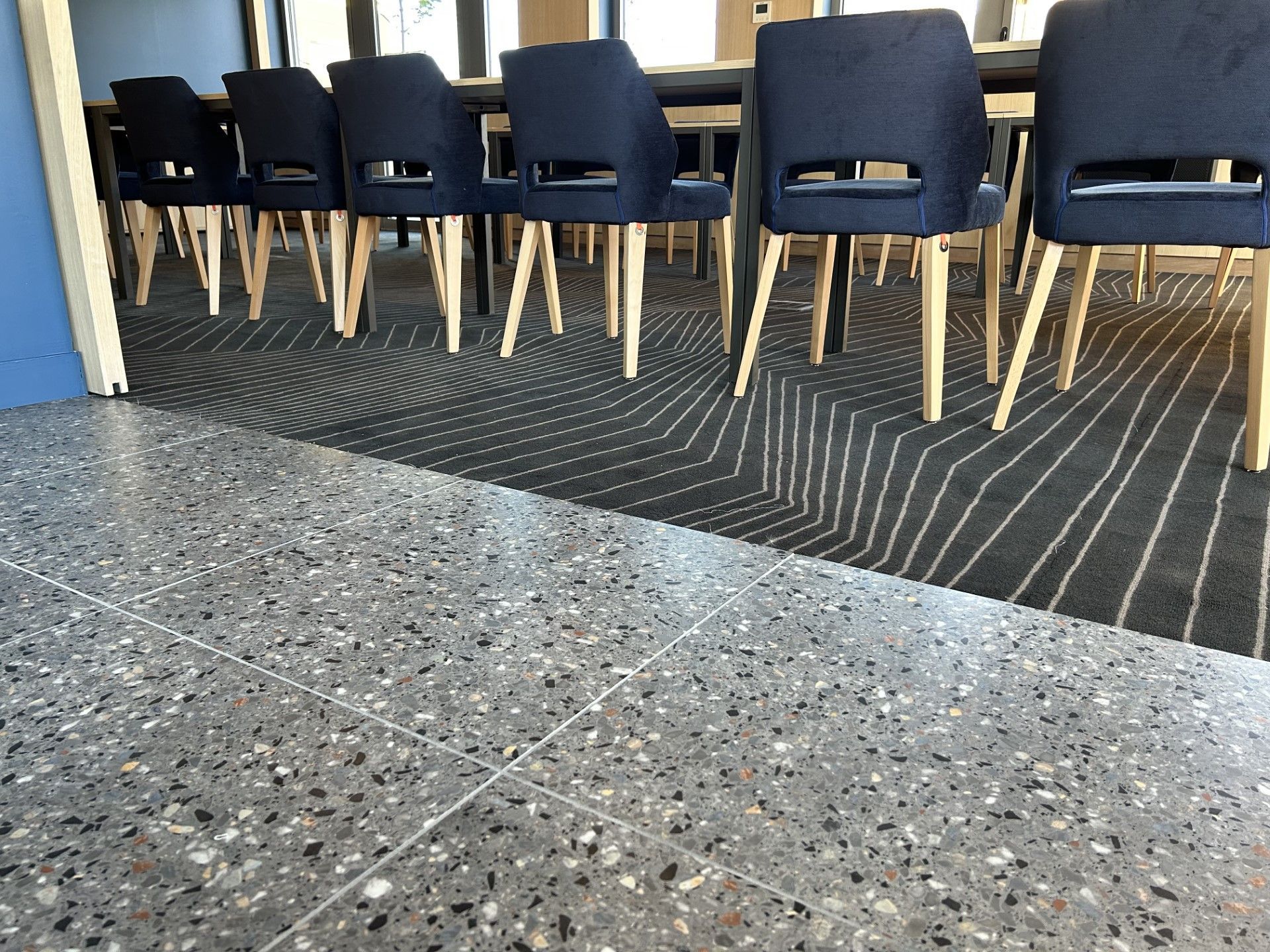 Un sol en terrazzo gris mène à une pièce avec une moquette à motifs, une table et des chaises bleues.
