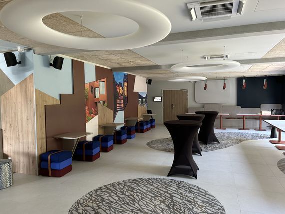 Intérieur d'un espace événementiel moderne avec une fresque colorée, des tables hautes, des tapis ronds et des sièges.
