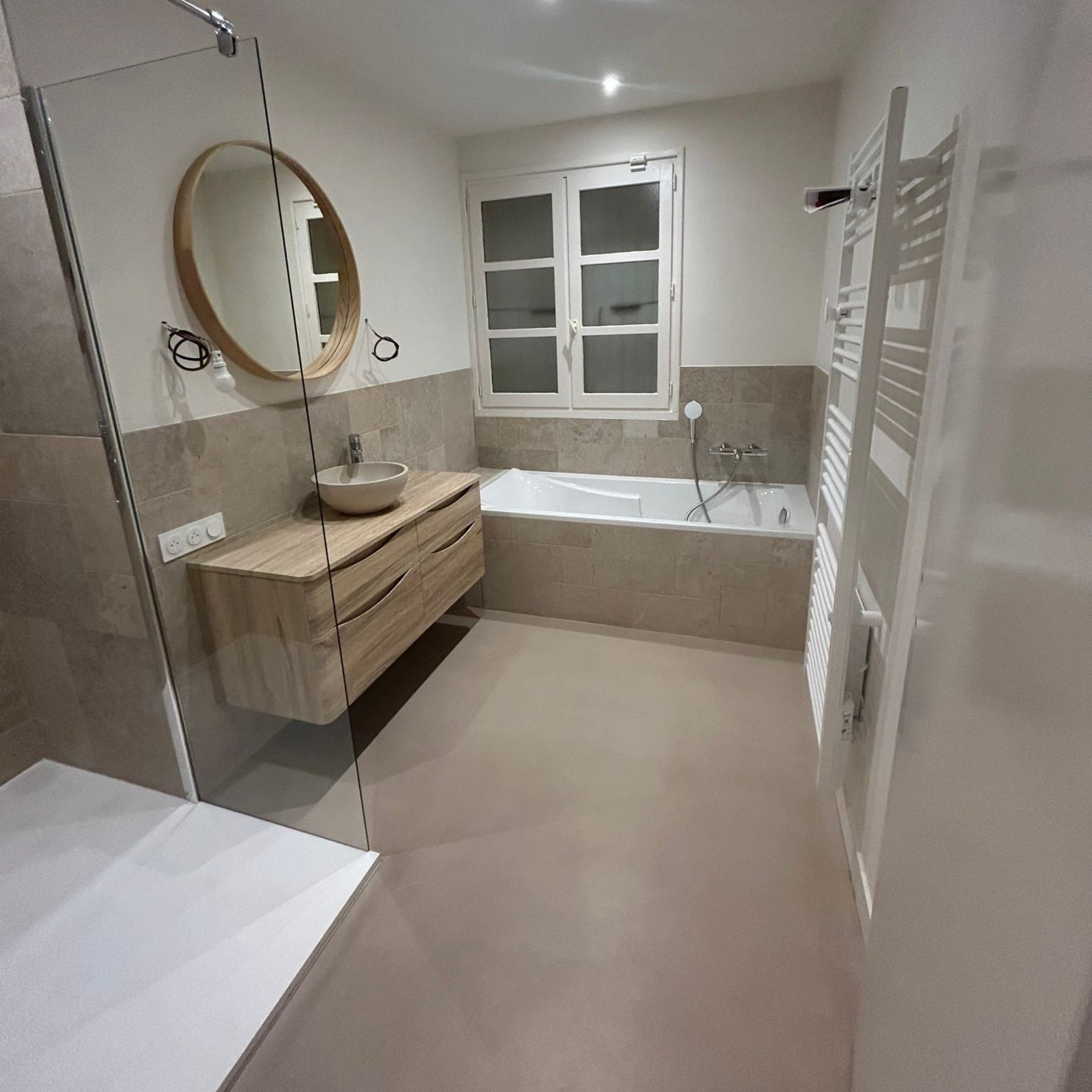 Salle de bains avec meuble-lavabo en bois, miroir rond, baignoire et fenêtre ; tons neutres.