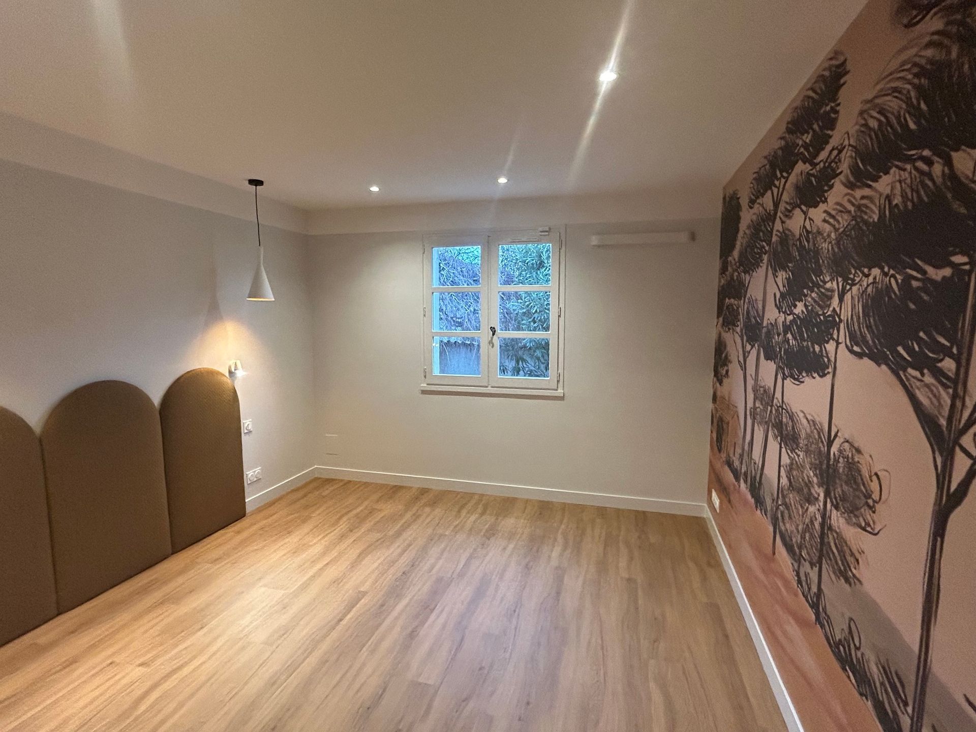 Chambre vide avec parquet clair, une fenêtre, une fresque murale sur un mur et une tête de lit.