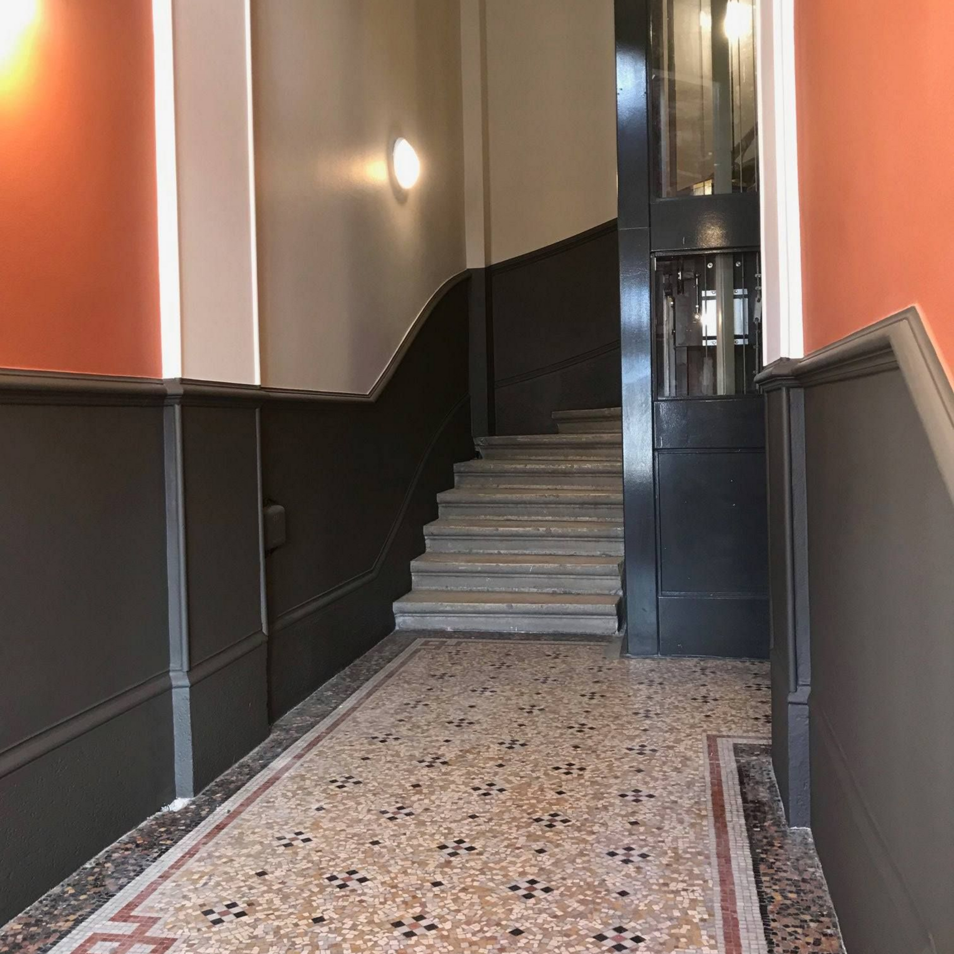 Entrée avec escalier et ascenseur. Murs orange/gris, sol à motifs.