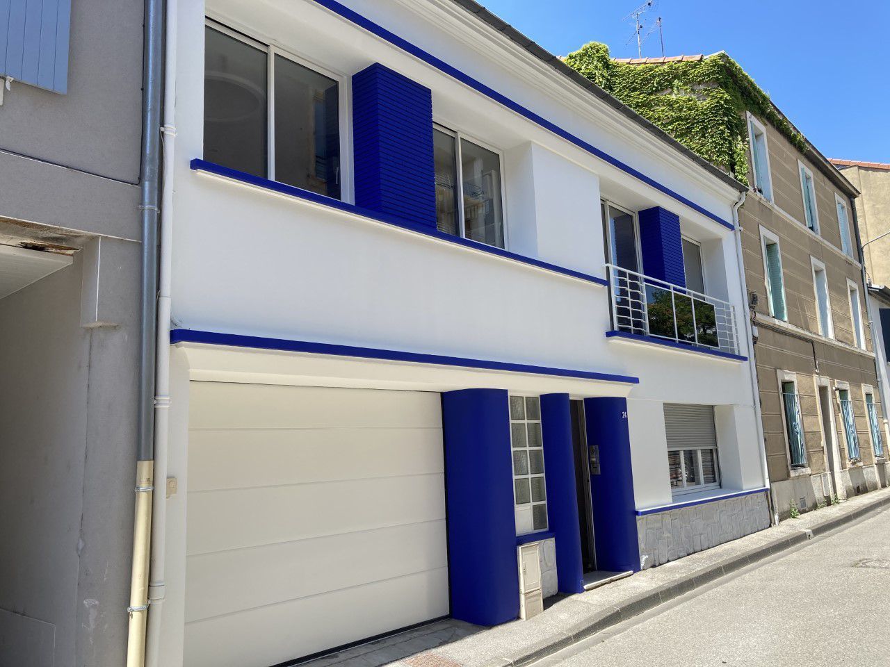 Maison blanche avec boiseries et porte de garage bleues, à deux étages, à côté d'un bâtiment en pierre.