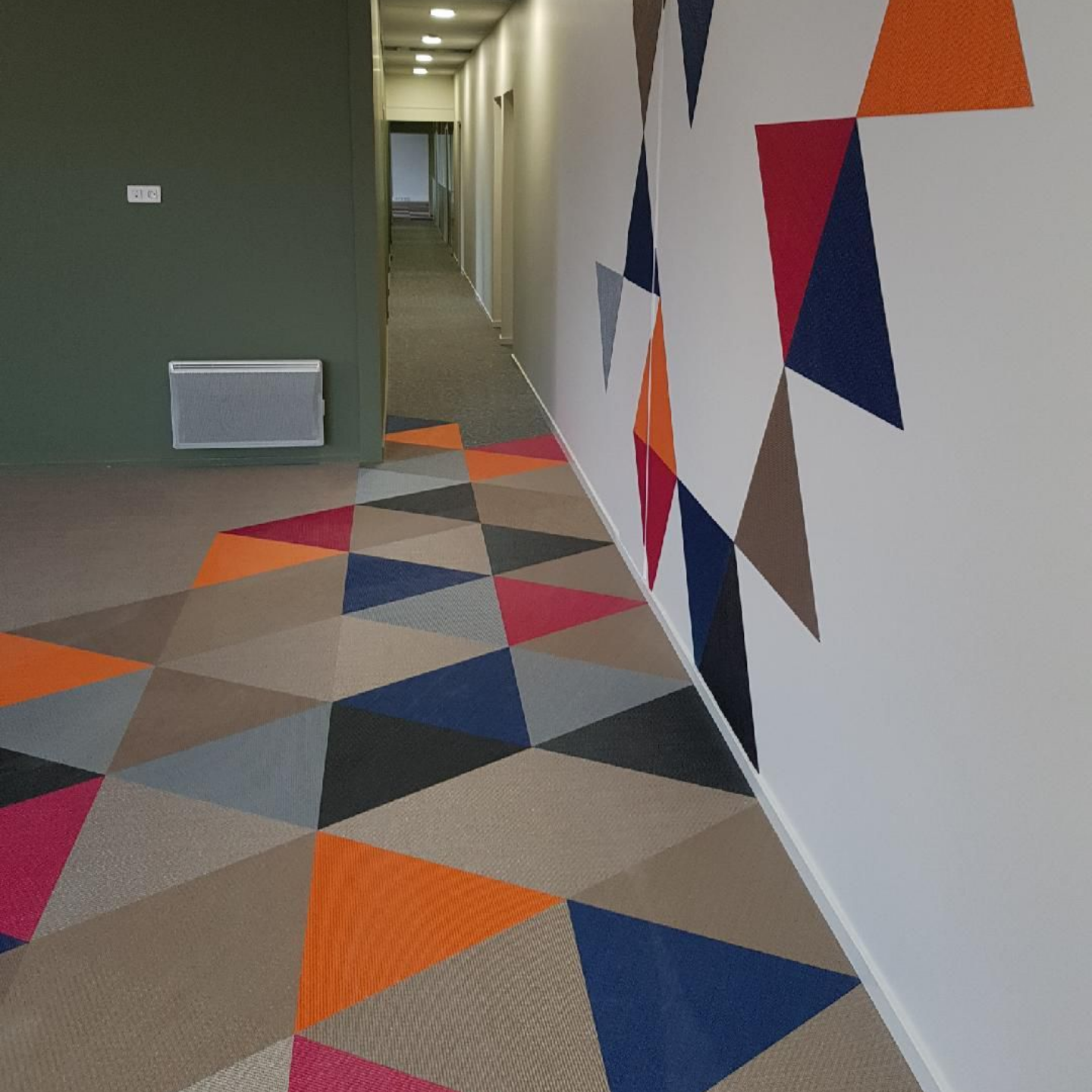 Couloir avec moquette et décoration murale géométriques. Les couleurs dominantes sont l'orange, le bleu, le rouge, le marron et le gris.