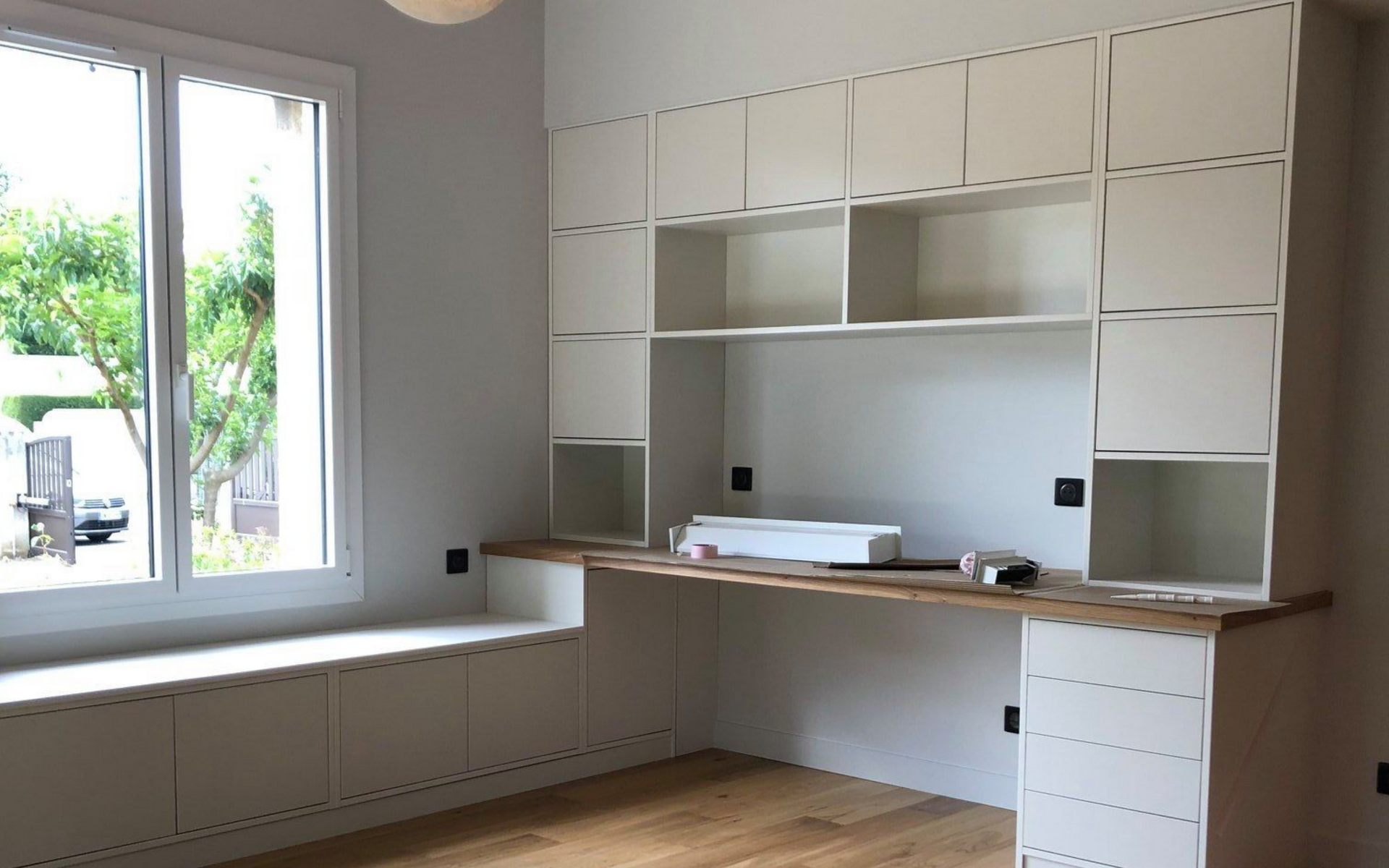 Bureau intégré avec placards blancs et bureau en bois, à côté d'une fenêtre avec banquette intégrée.