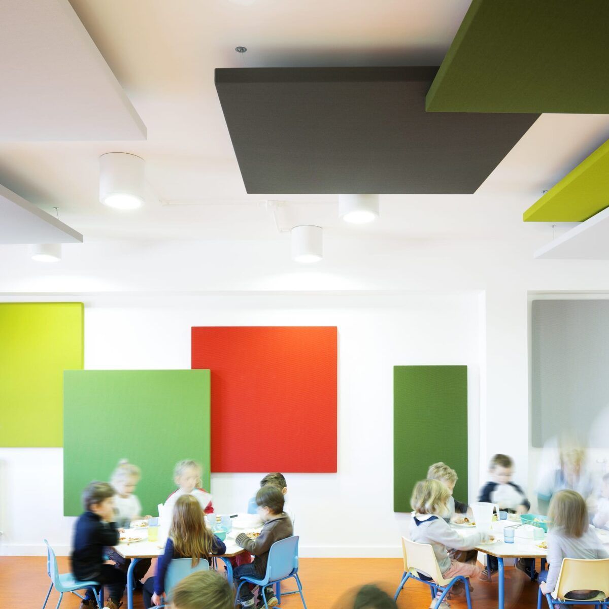 Salle de classe avec panneaux d'insonorisation colorés ; enfants assis à des tables, silhouettes floues.
