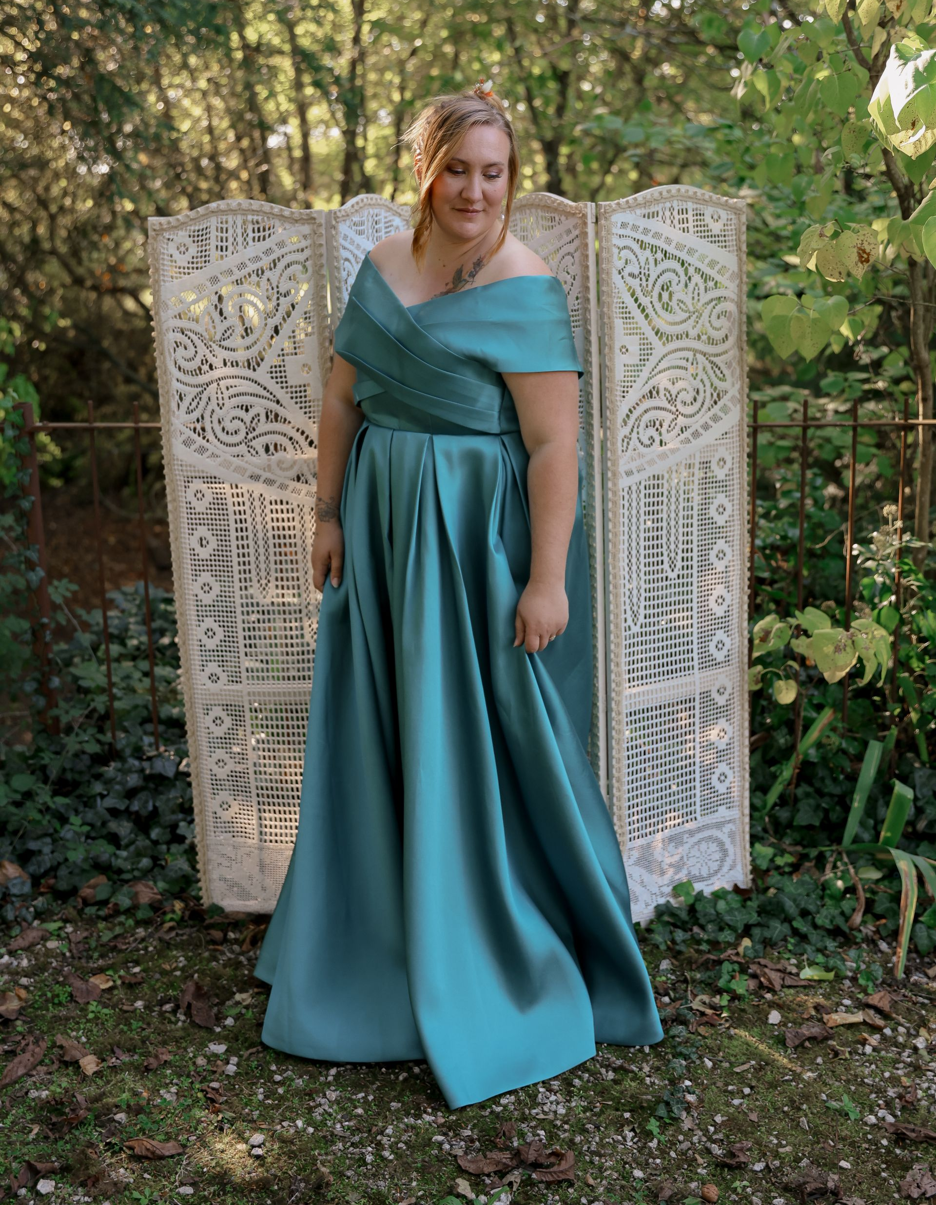 Une femme vêtue d'une robe turquoise à épaules dénudées se tient à l'extérieur, devant un paravent décoratif, souriante.