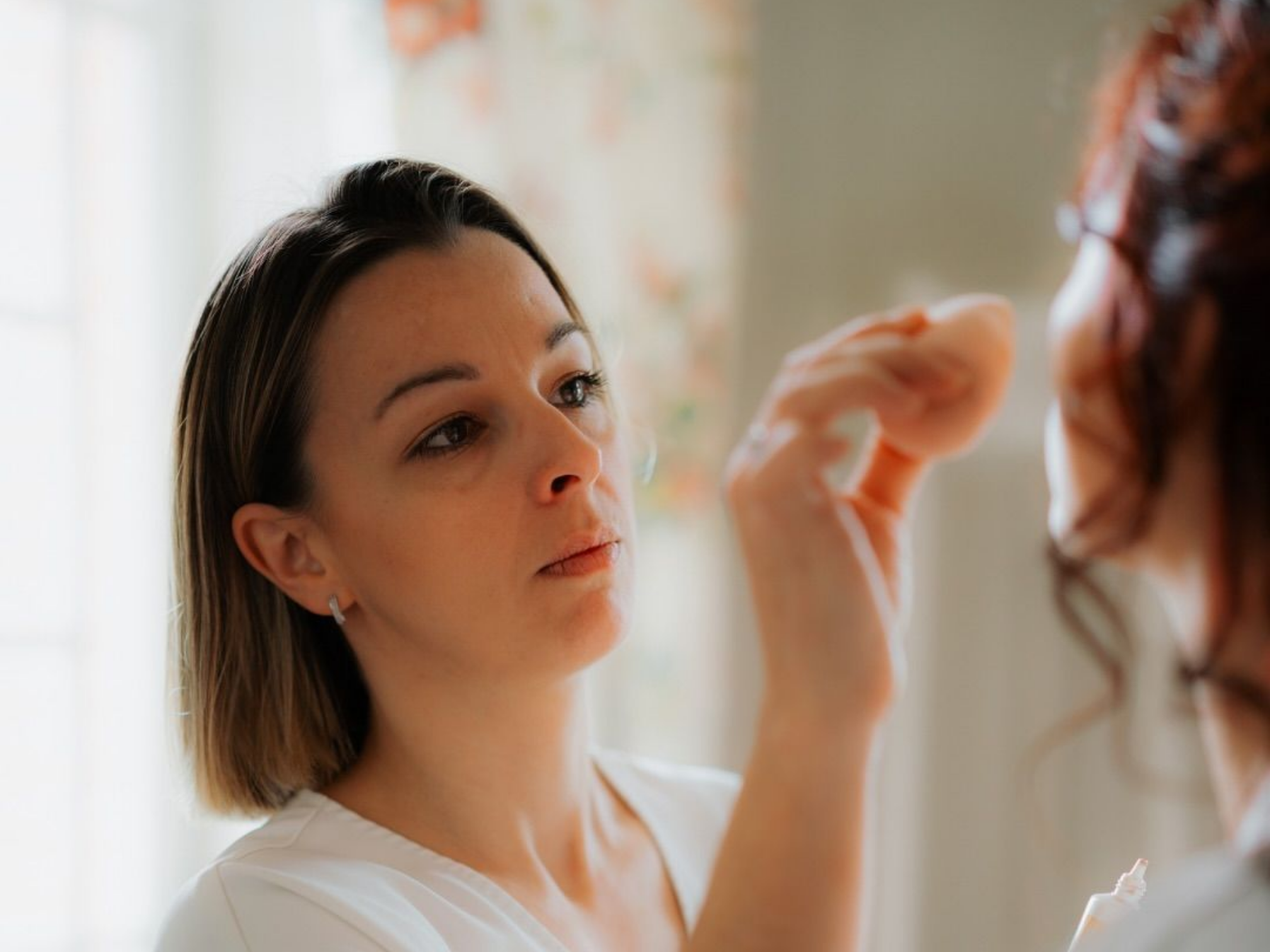 Une femme applique du maquillage sur le visage d'une autre femme avec une éponge dans un cadre intérieur lumineux.