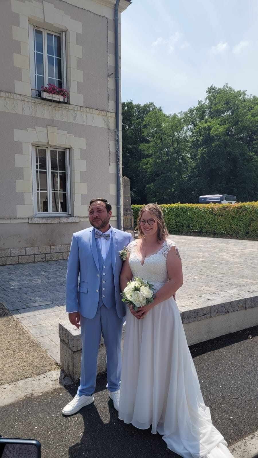 Les mariés posent devant un bâtiment. Le marié porte un costume bleu clair. La mariée porte une robe blanche et tient un bouquet de fleurs.