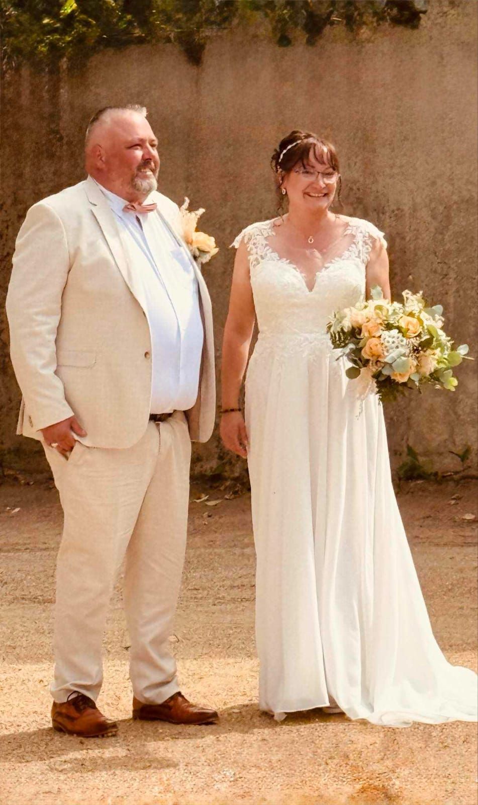 Les mariés posent en extérieur. La mariée, en robe blanche, tient son bouquet. Le marié porte un costume.