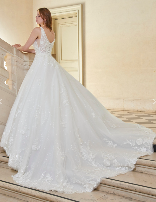 La mariée en robe blanche ornée de perles regarde par-dessus son épaule. Le marié en costume sourit derrière elle. Décor extérieur.