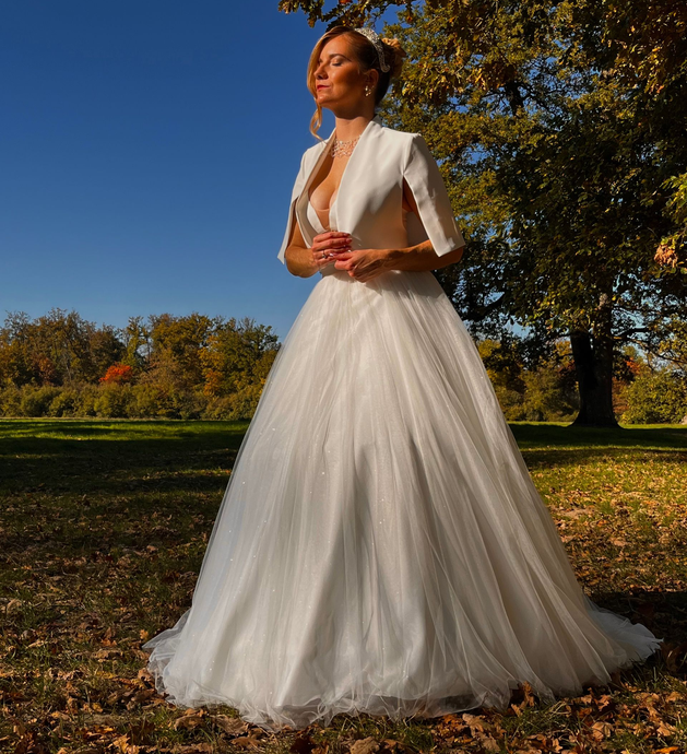 Mariée en robe blanche à épaules dénudées tenant un bouquet, souriant, regardant vers le haut avec les yeux fermés sur un fond neutre.