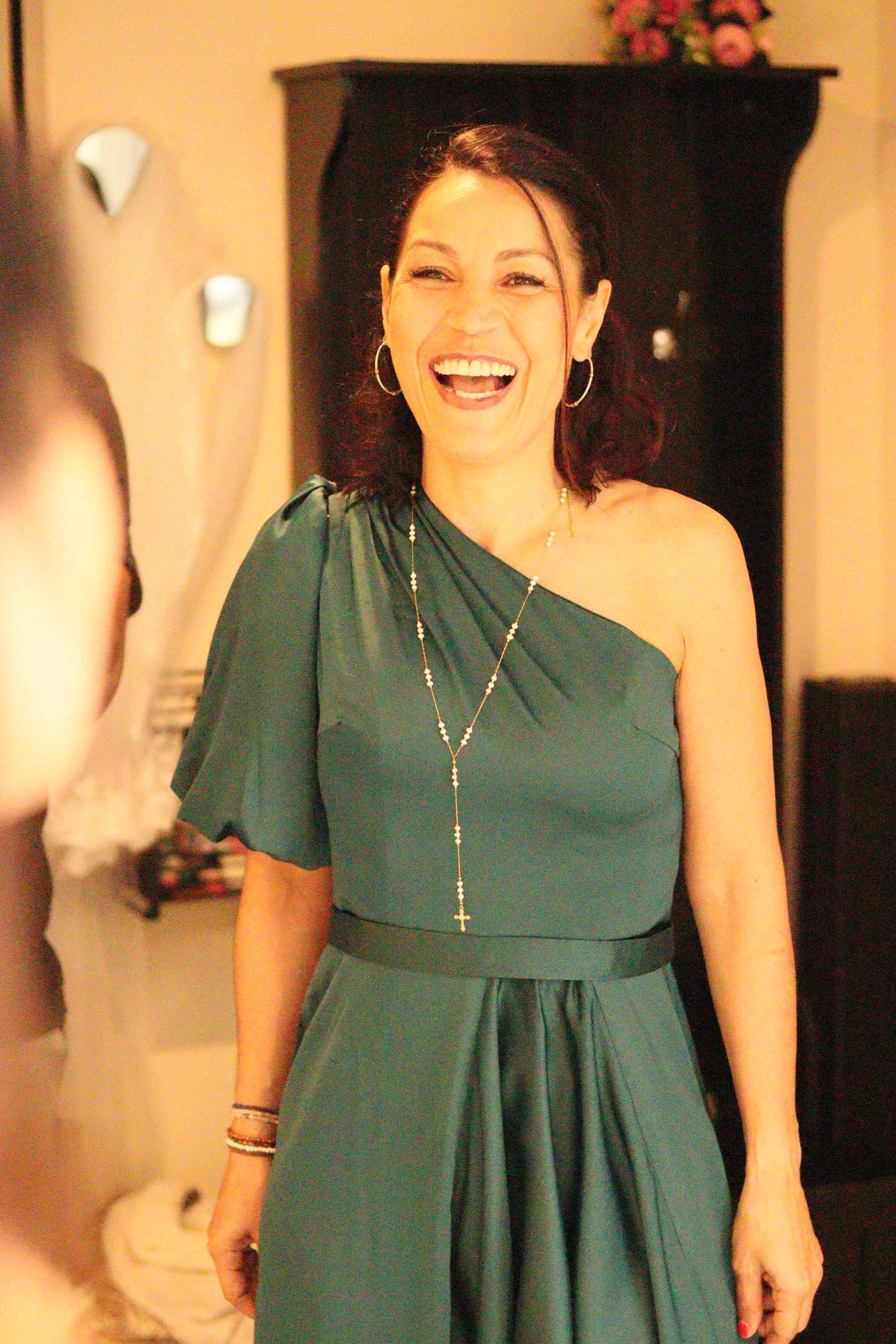 Une femme vêtue d'une robe asymétrique turquoise rit à l'intérieur. Elle porte un long collier et a les cheveux foncés.