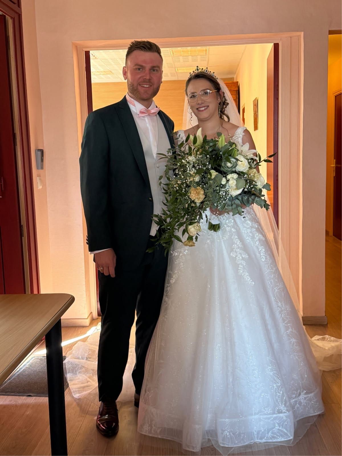 Les mariés posent sur le seuil d'une porte : l'homme en costume, la femme en robe blanche.