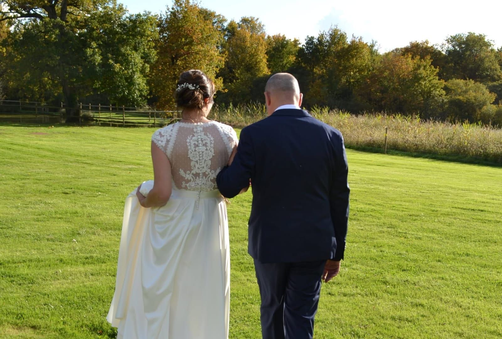 Une mariée en robe blanche marche avec un homme en costume à travers un champ.