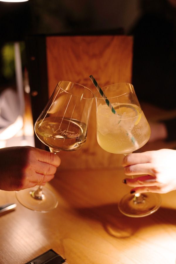 Zwei Hände stoßen mit Wein- und Cocktailgläsern an einem warm beleuchteten Holztisch an.