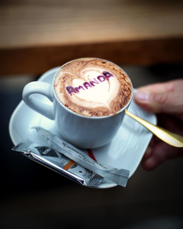 Eine Tasse Cappuccino mit herzförmiger Schaumverzierung und dem Schriftzug „AMANDA“ darauf, serviert auf einem Unterteller mit Zuckerpäckchen.