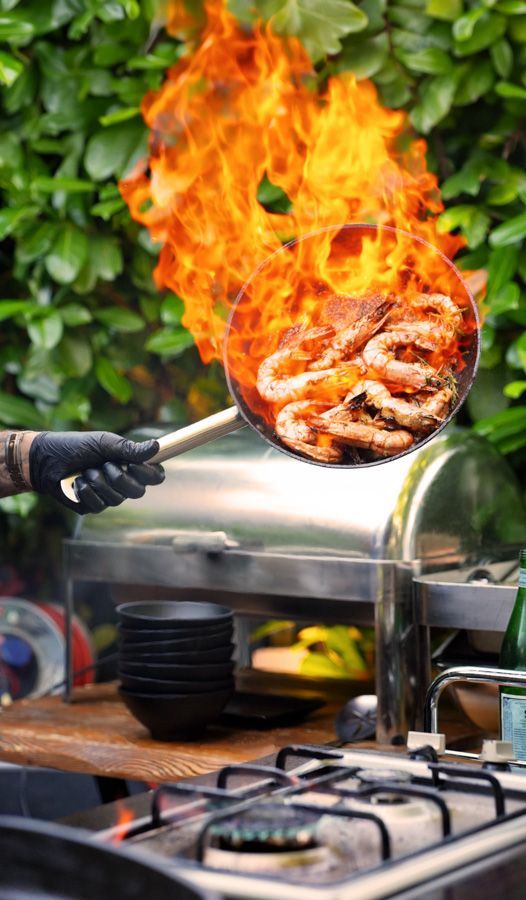 Gegrilltes Fleisch, das auf einem Grill im Freien in Flammen steht, mit einer behandschuhten Hand, die eine Grillzange benutzt.
