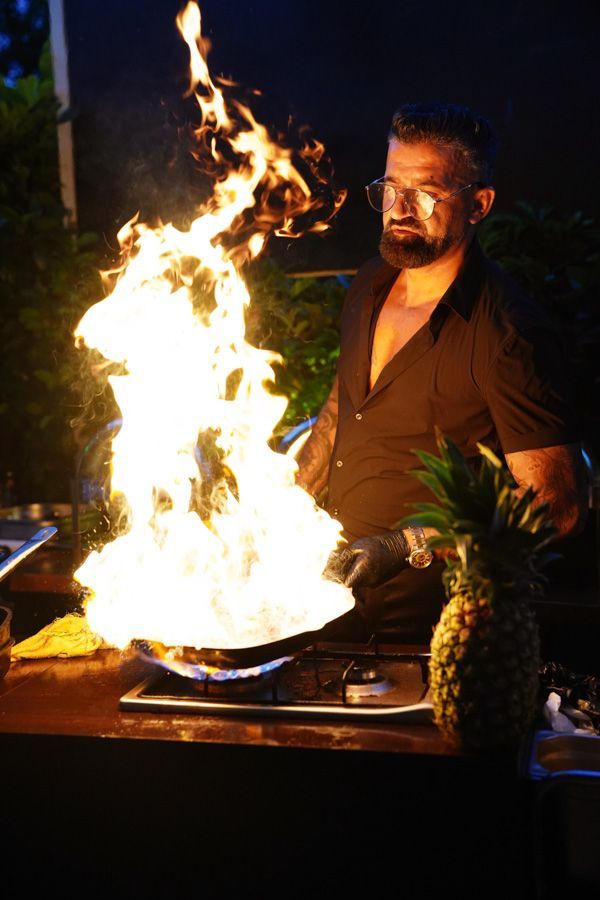 Ein Koch bereitet nachts Speisen über einer brennenden Pfanne zu; helle Flammen lodern, und auf dem Tisch steht eine Ananas.