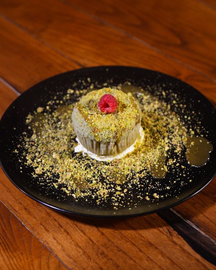 Kleines Dessert mit einer Himbeere auf einem schwarzen Teller, bestreut mit Krümeln, auf einem Holztisch.