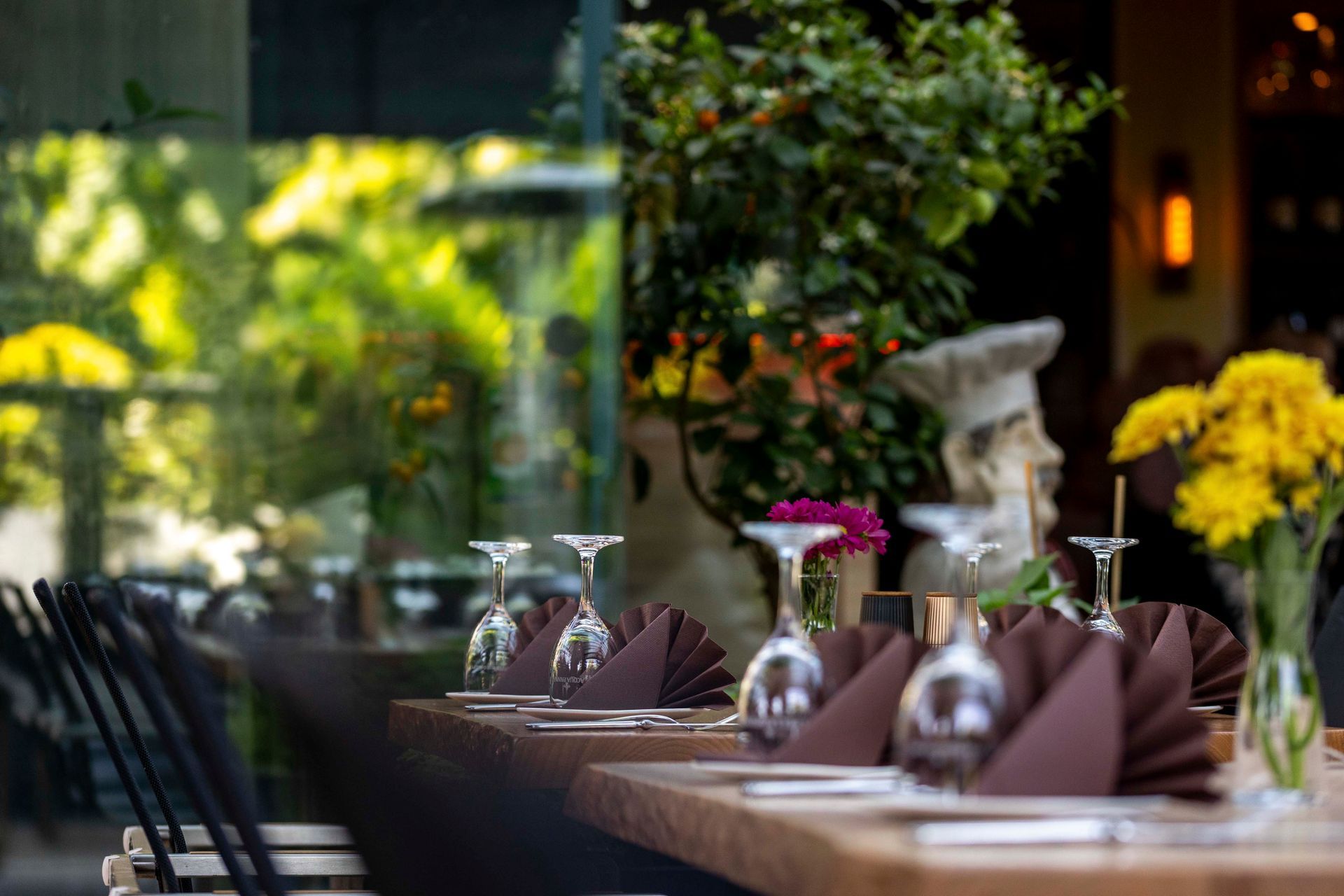 Gedeckte Restauranttische, durch ein Glasfenster gesehen, mit braunen Servietten, Weingläsern und Blumen. Im Hintergrund ist üppiges Grün zu sehen.