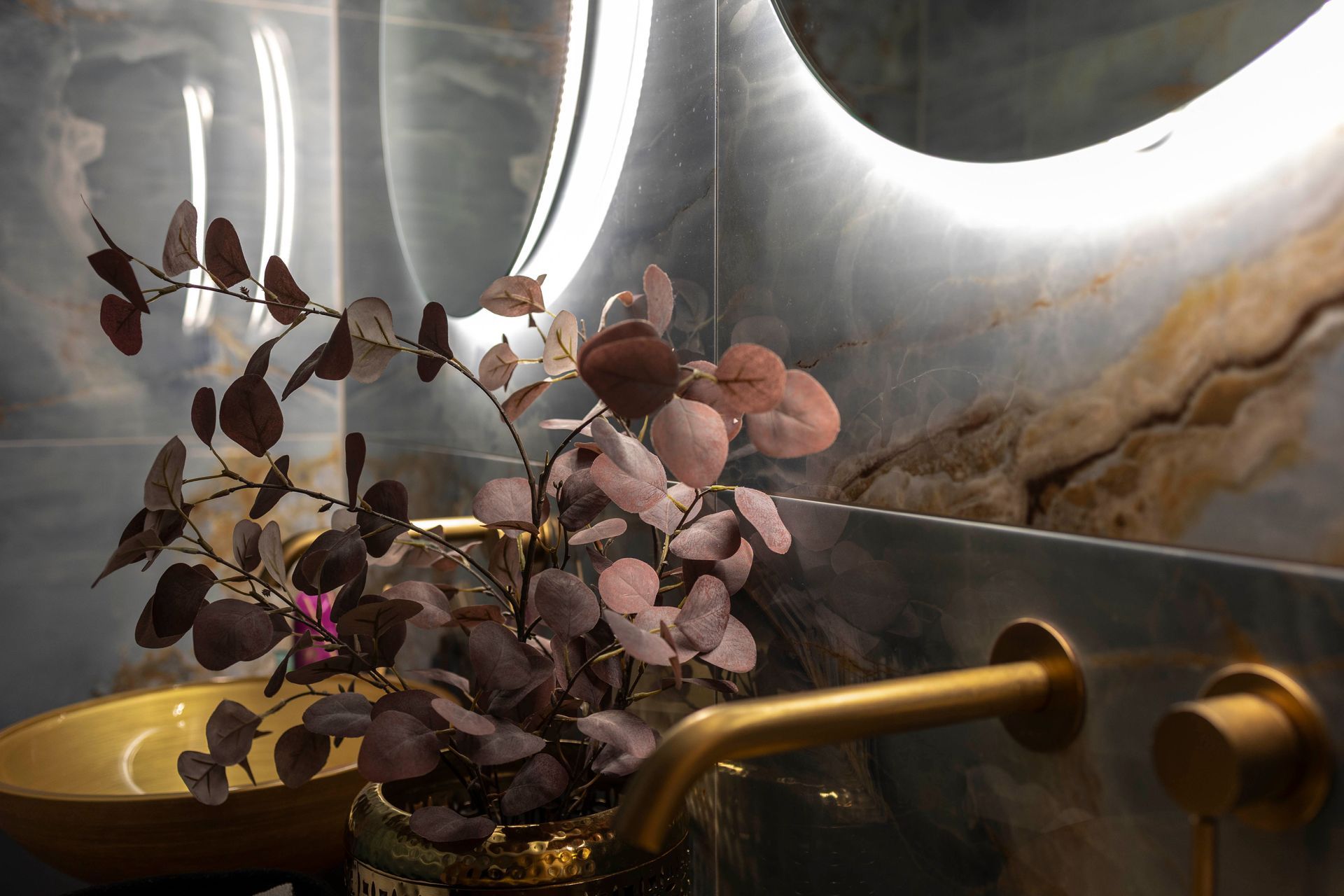 Nahaufnahme eines modernen Badezimmers mit goldenem Waschbecken, Wasserhahn und Vase mit rötlich-violetten Blättern. Im Hintergrund befindet sich ein runder, hinterleuchteter Spiegel.