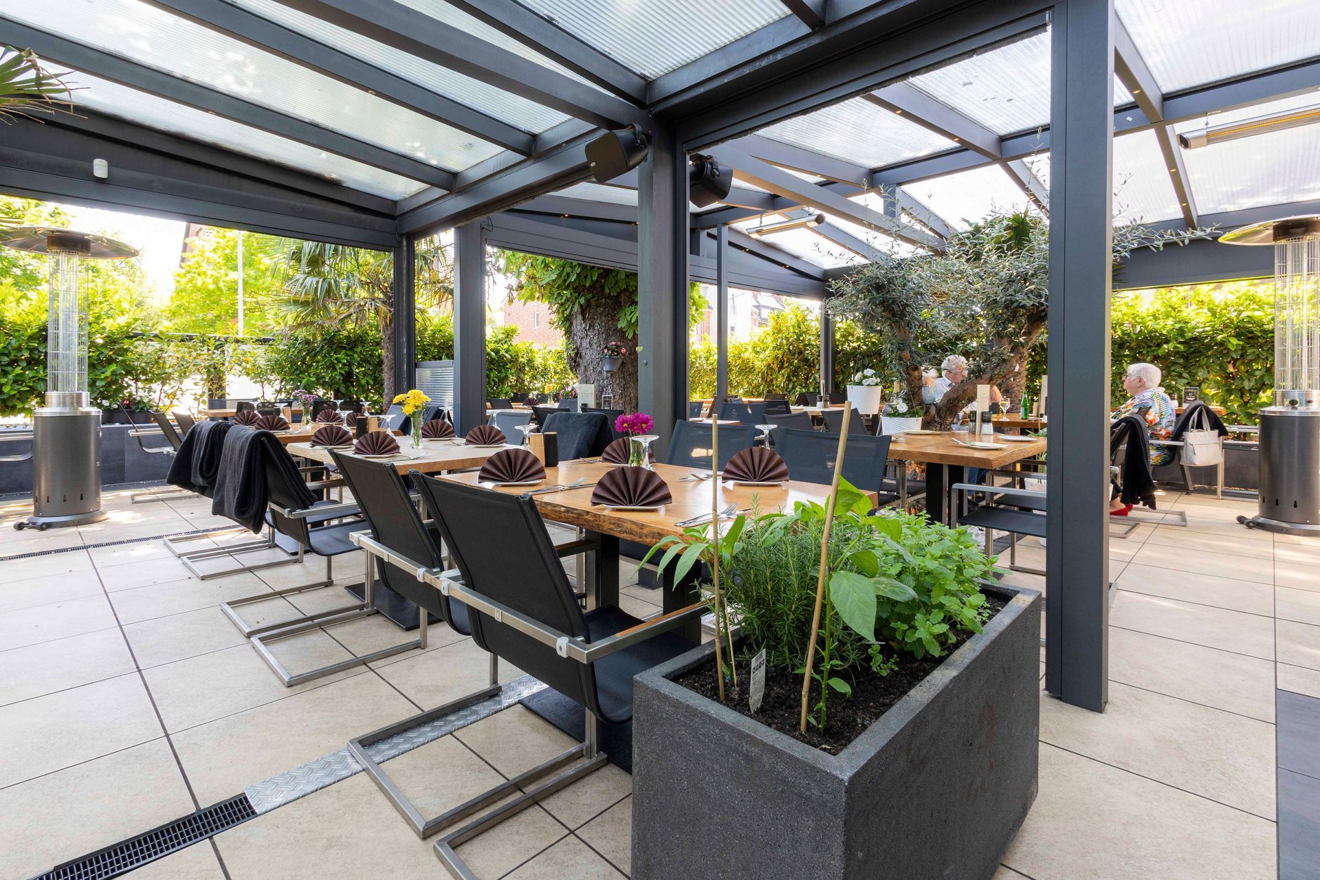 Essbereich im Freien mit Tischen und Stühlen unter einem Baldachin. Die Terrasse verfügt über Heizstrahler, einen Pflanzkasten und einige Sitzgelegenheiten.