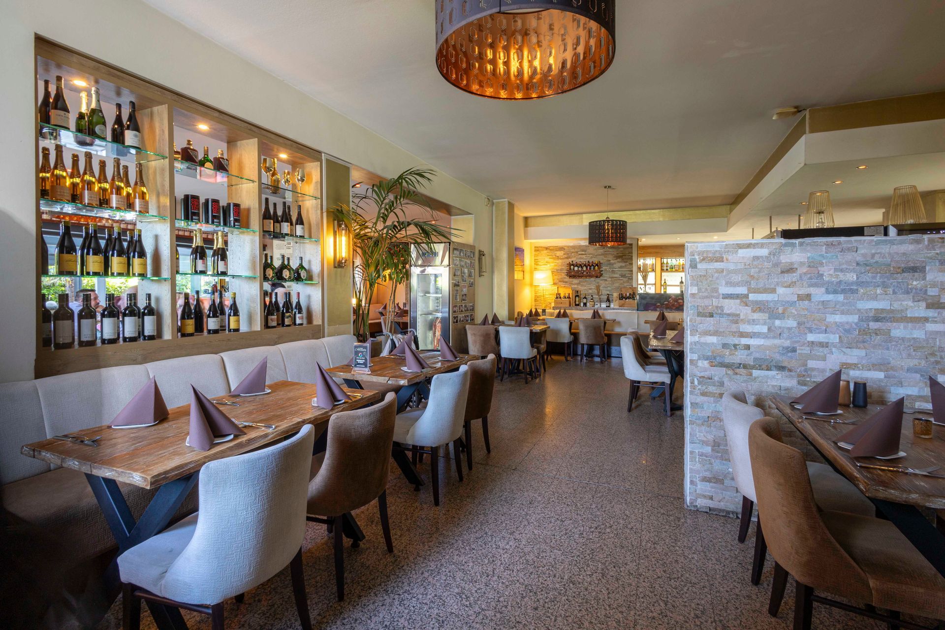 Restaurant-Innenraum mit gedeckten Tischen, einer Wand aus Weinflaschen und Dekor in neutralen Farbtönen.