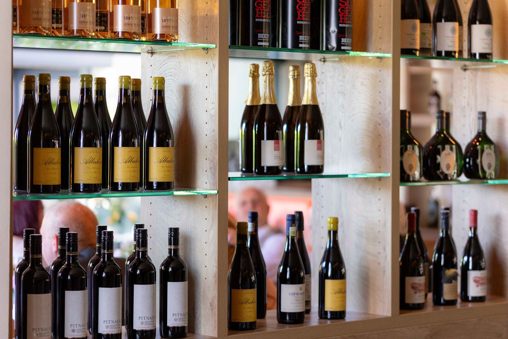 Weinflaschen in Ausstellungsregalen in einem Weinladen, mit verschiedenen Etiketten und Flaschenformen. Verschwommene Figuren im Hintergrund.