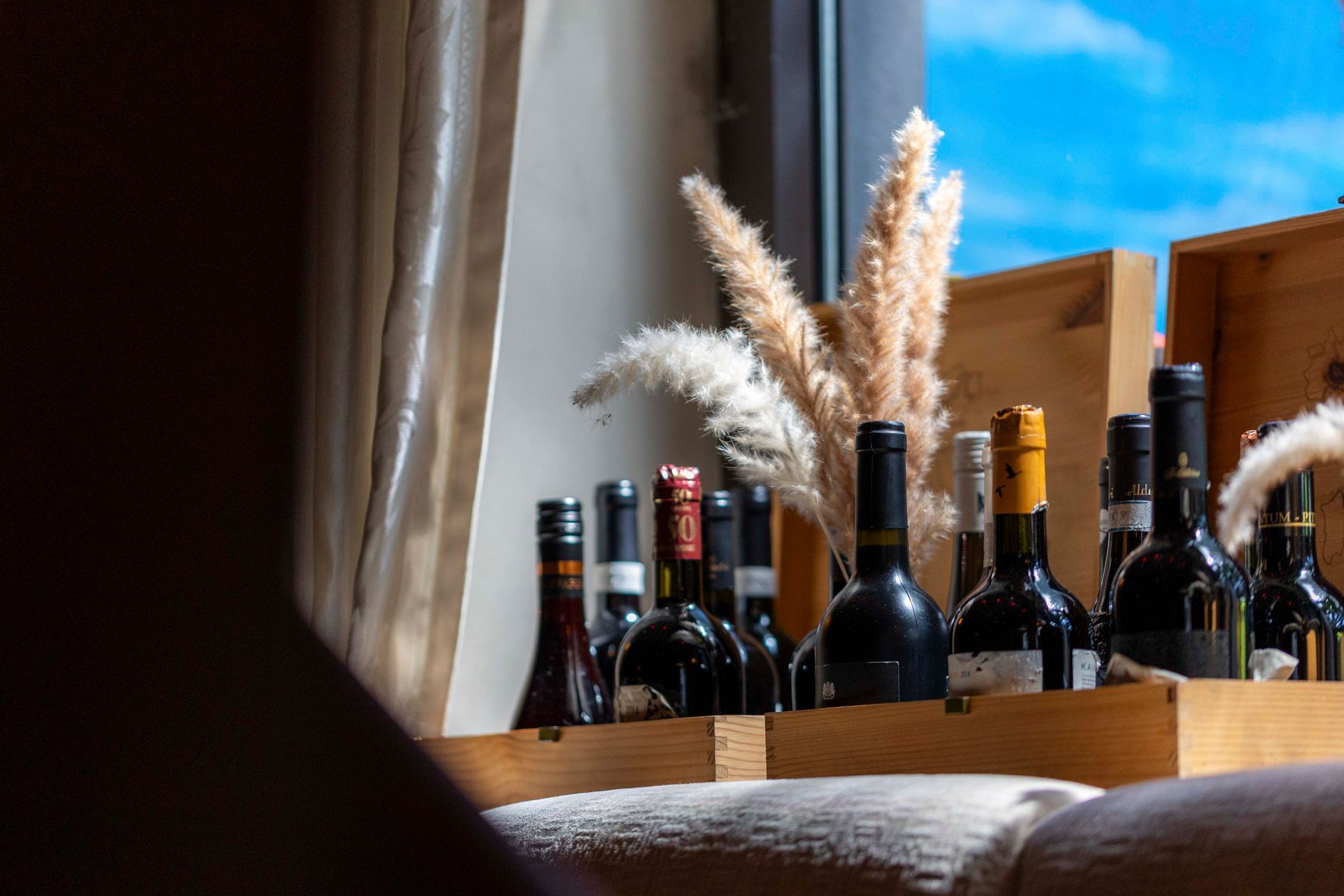 Weinflaschen in Holzkisten, mit dekorativen getrockneten Gräsern, neben einem Fenster mit strahlend blauem Himmel.