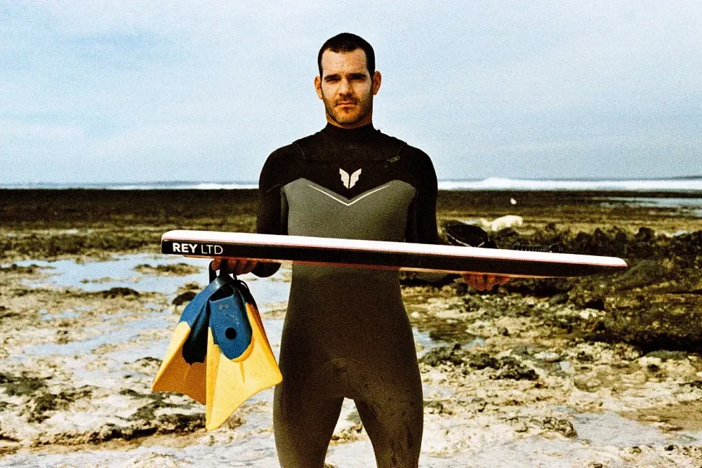 Un hombre con traje de neopreno sostiene una tabla de surf que dice