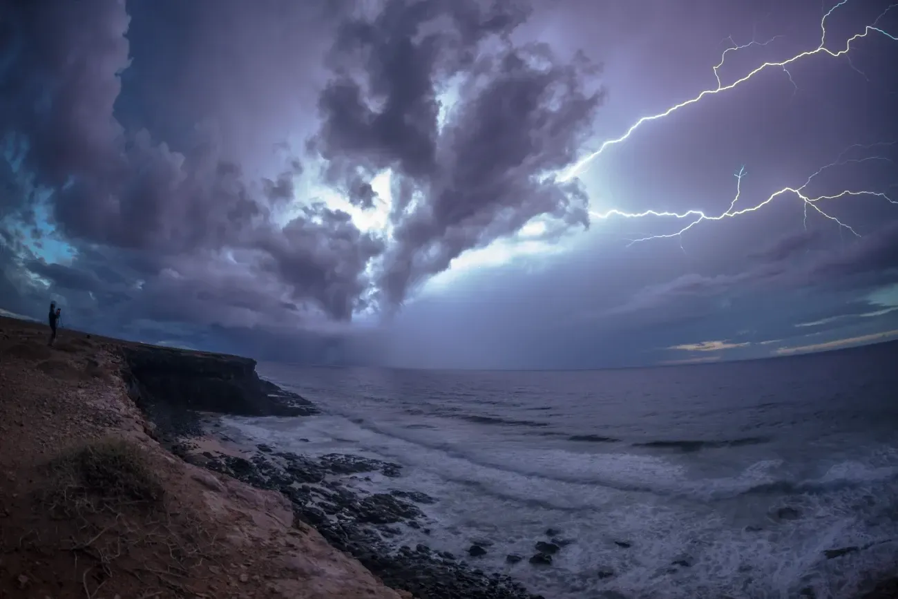 Una persona está parada en un acantilado con vista al océano durante una tormenta eléctrica.