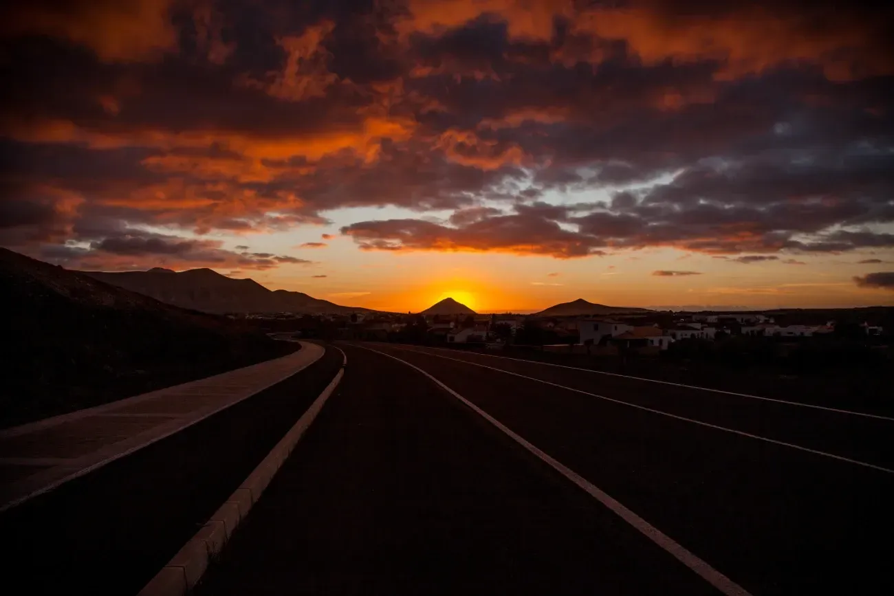 Una carretera con un atardecer al fondo y una montaña a lo lejos.
