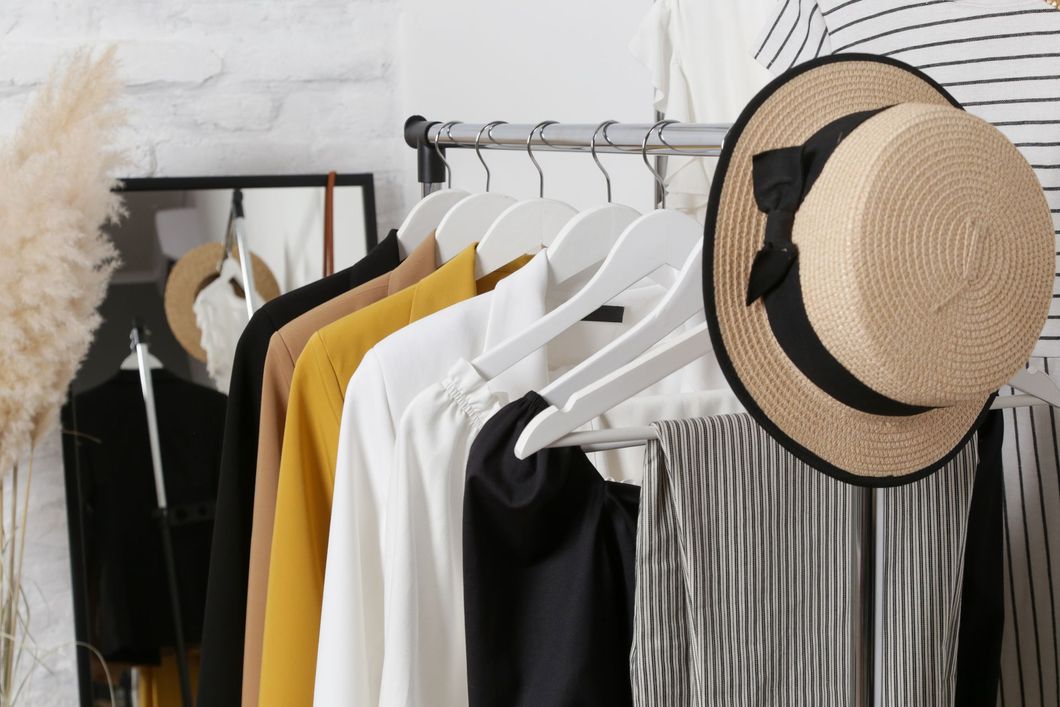 Un portant à vêtements avec des vêtements blancs, jaunes et noirs, des miroirs encadrés et un chapeau de paille dans une pièce lumineuse.